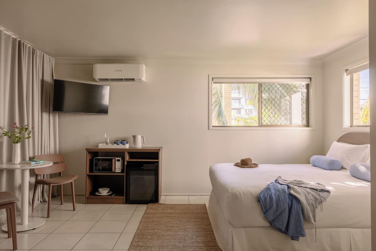 Une chambre d’hôtel avec un lit, un téléviseur fixé au mur, une petite table avec des chaises, une bibliothèque et une fenêtre donnant sur des immeubles.