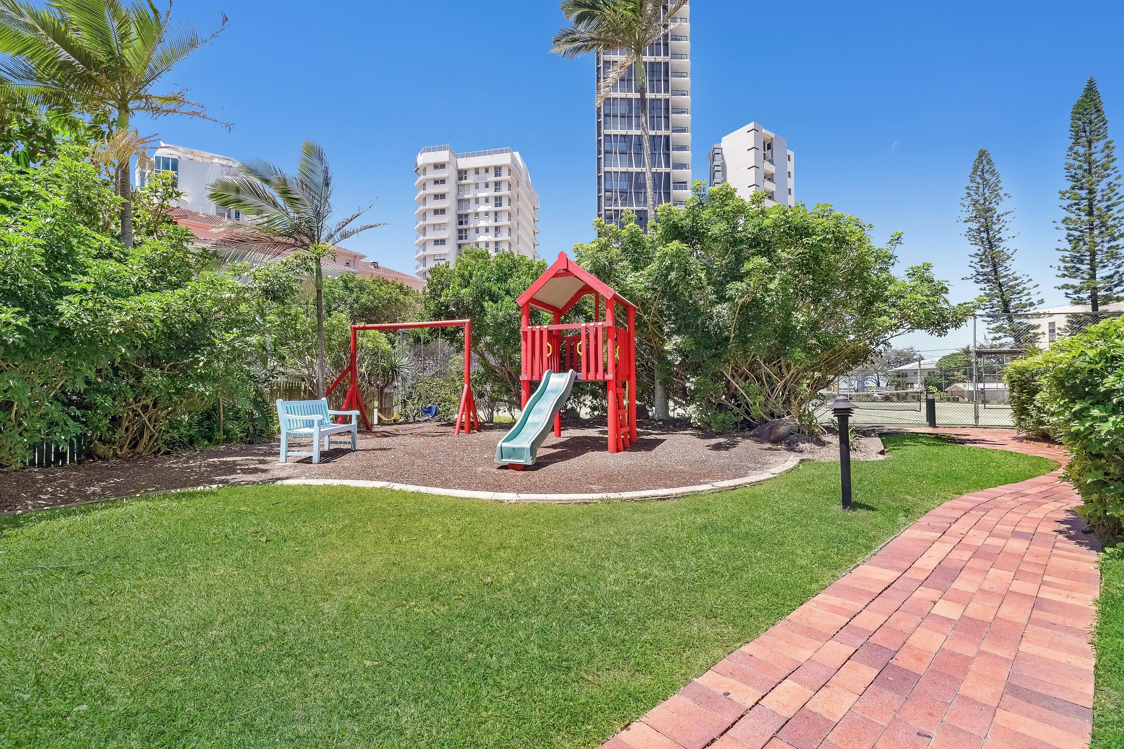A playground with red structures and a slide, surrounded by greenery and a brick pathway.