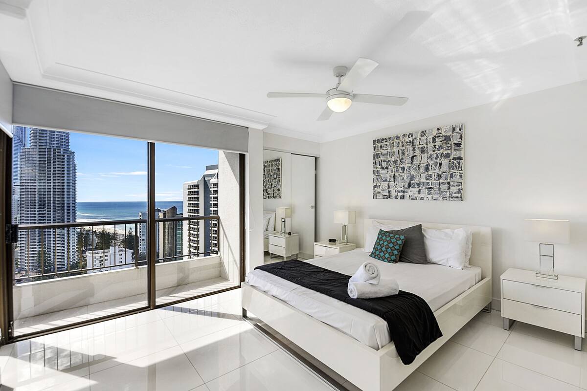 Habitación de hotel moderna con una cama grande, ventilador de techo, vista al océano y al paisaje urbano.