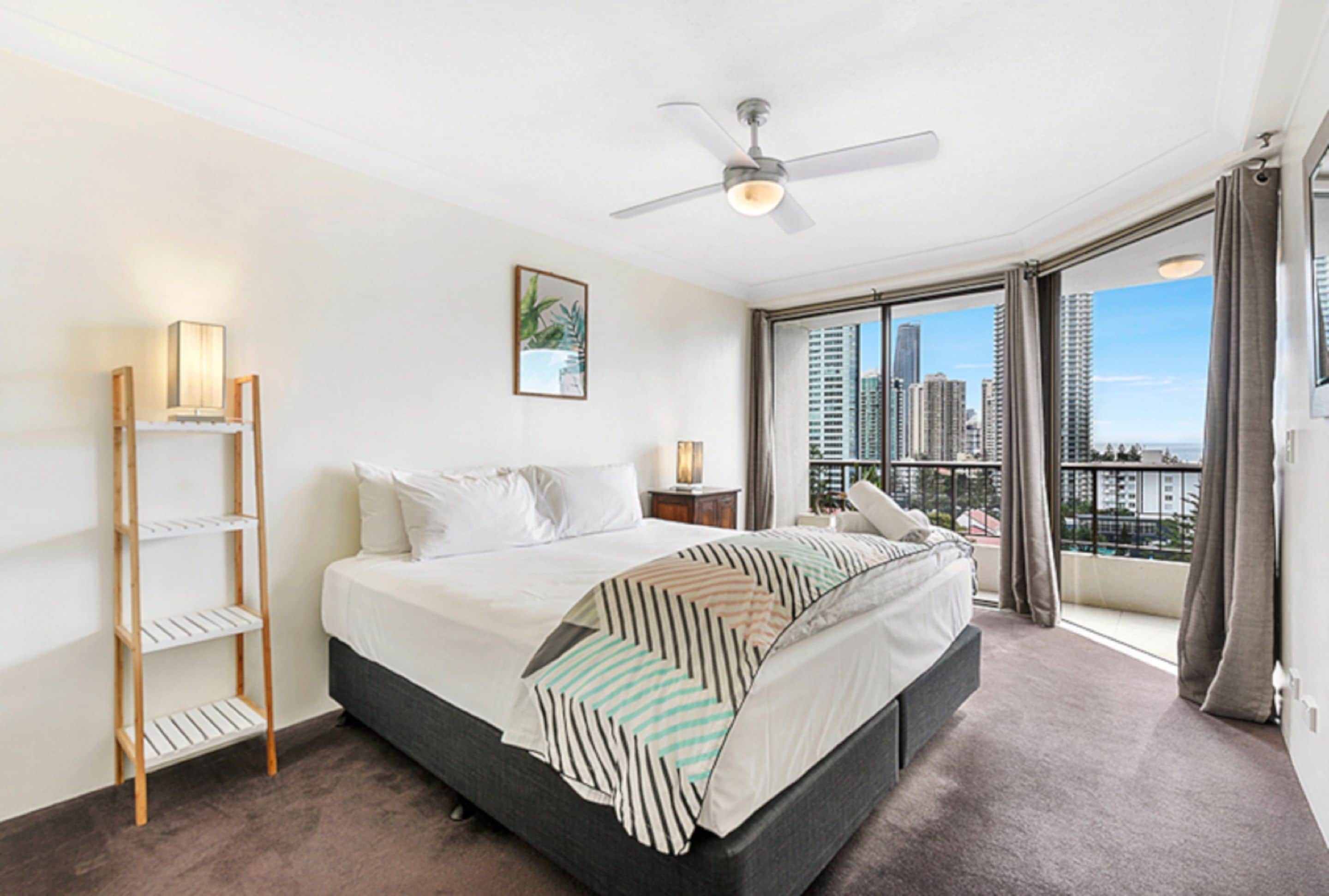 A hotel room with a bed, a ceiling fan, a city view, and a balcony.