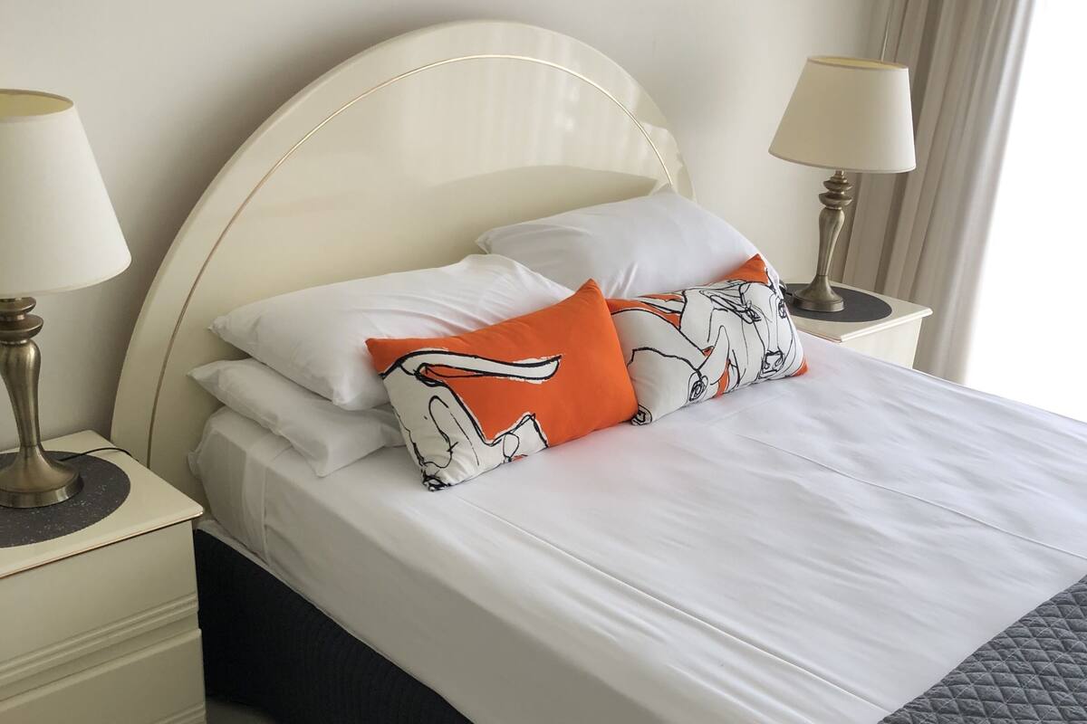Una cama bien hecha con sábanas blancas y dos almohadas, una naranja y otra con estampado blanco y negro. Hay dos mesitas de noche con lámparas, y una ventana con cortinas.