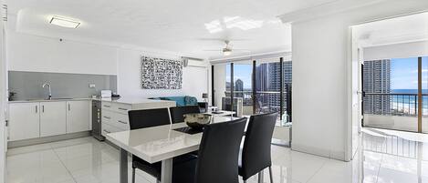 Une cuisine moderne avec des meubles blancs, une table à manger avec des chaises et une vue sur la ville grâce à de grandes fenêtres.