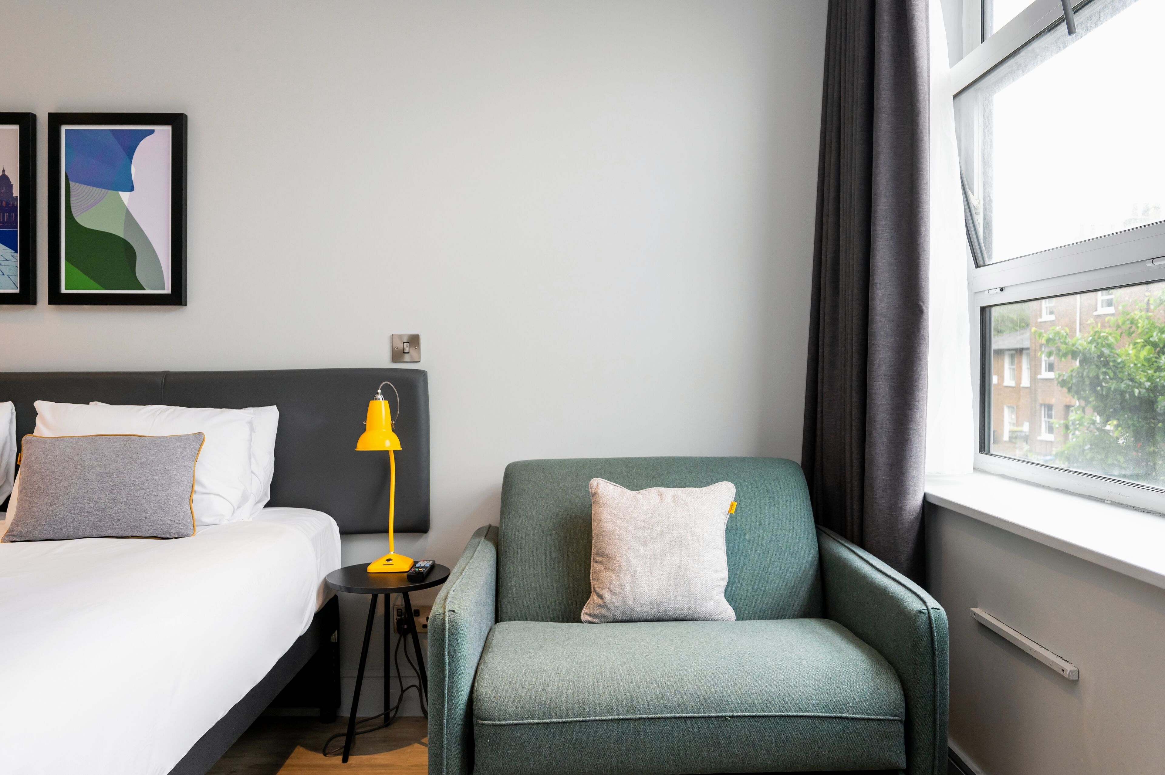 A hotel room with a bed, a chair, a nightstand, a lamp, and a window with a view of buildings.