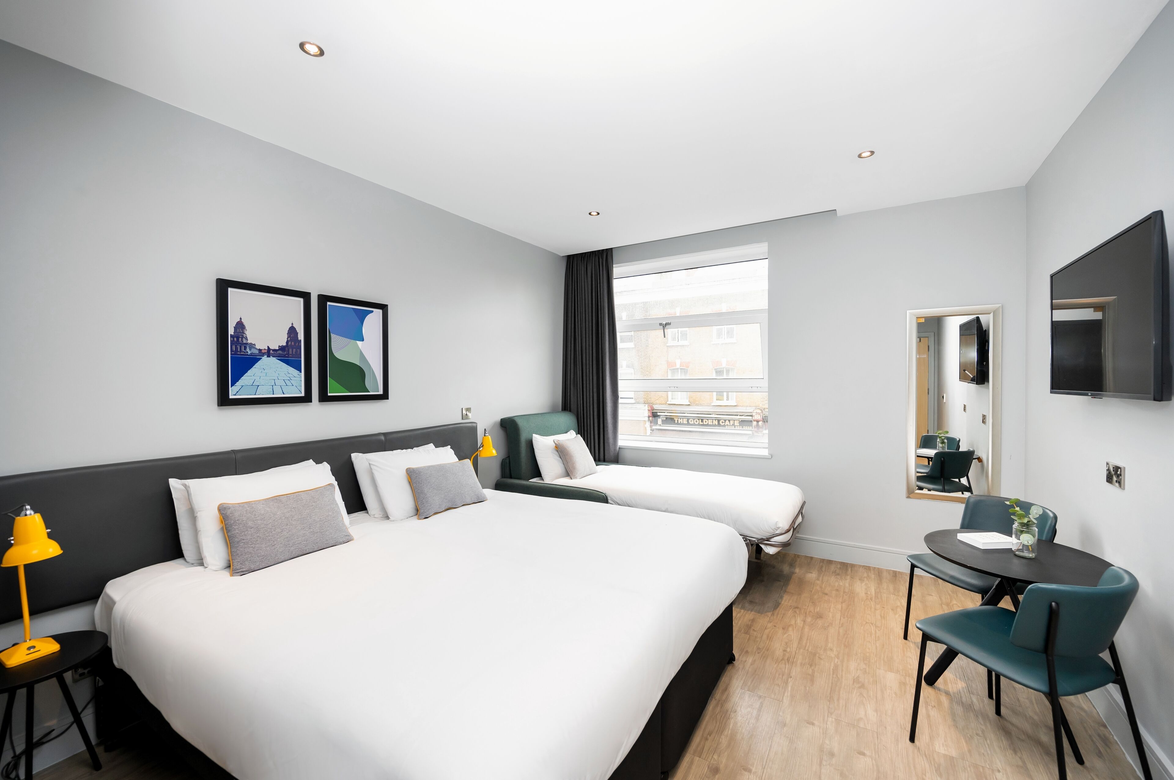 A hotel room with two beds, a desk, a chair, a TV, and a window with a view of buildings.