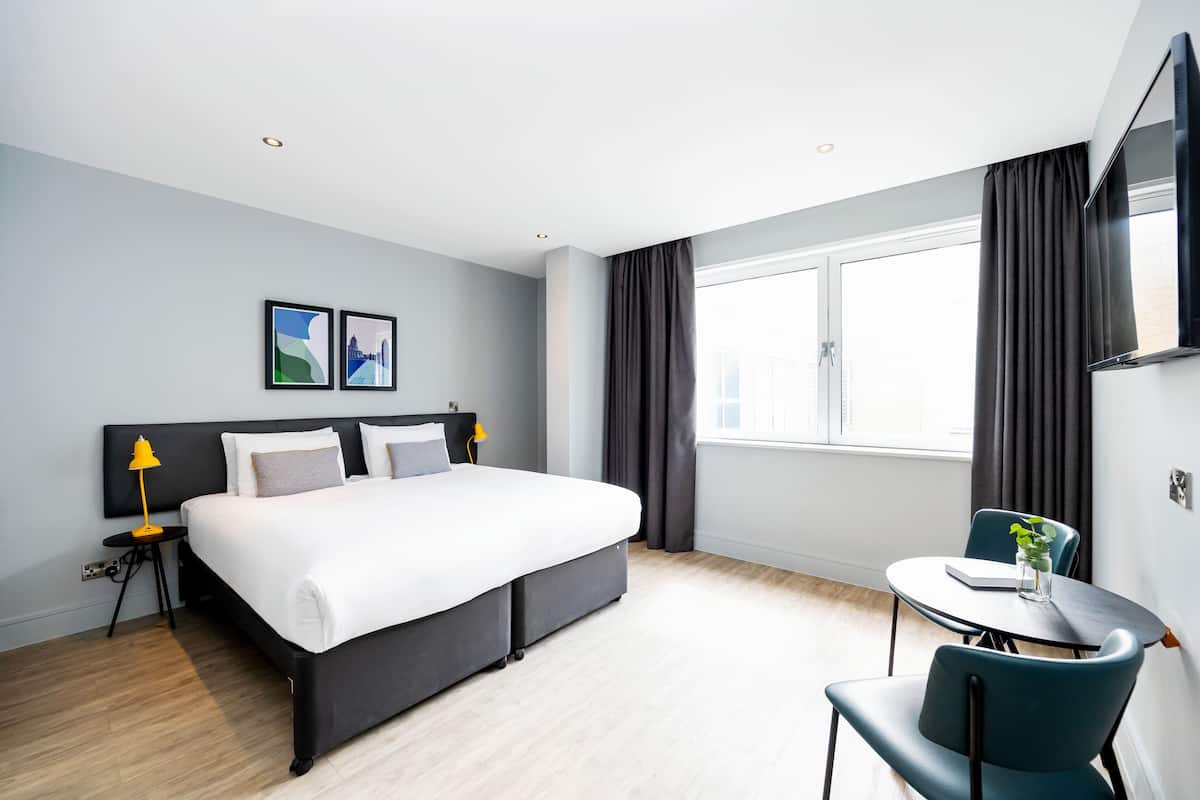 Une chambre d’hôtel moderne avec un grand lit, une petite table ronde avec une chaise, un téléviseur fixé au mur et une fenêtre avec des rideaux.