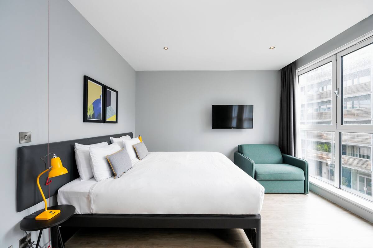 Une chambre d’hôtel moderne dotée d’un grand lit, d’un bureau avec une lampe, d’un téléviseur fixé au mur et offrant une vue sur la ville grâce à de grandes fenêtres.