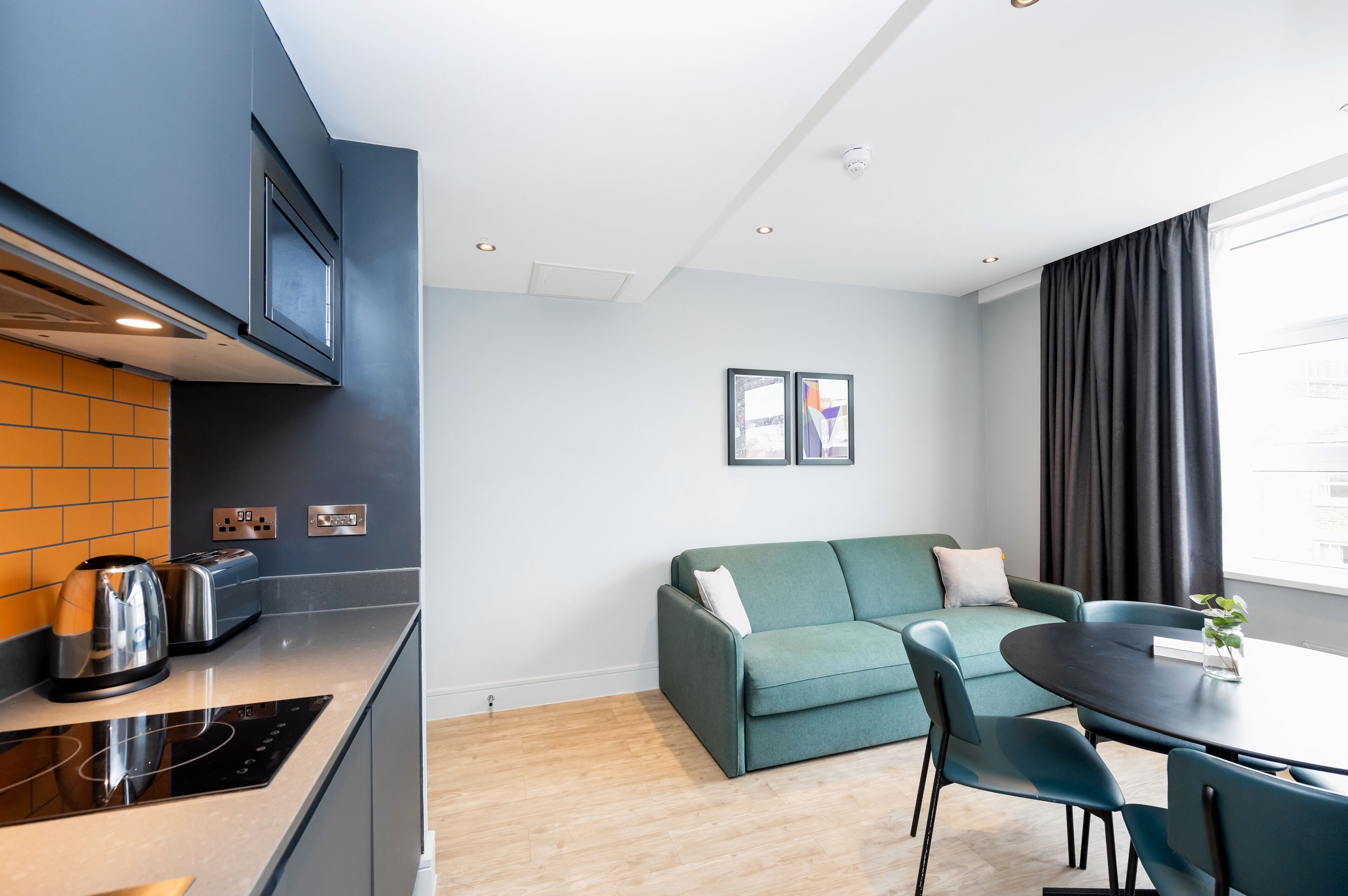 A modern kitchen with a built-in stove, a toaster, and a kettle. There is a dining area with a table and chairs, and a sofa. The room has a large window with curtains.