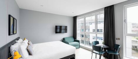 A modern hotel room with a large bed, a desk with two chairs, a TV on the wall, and large windows with curtains.