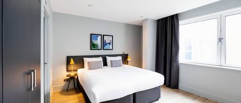 A modern hotel room with a large bed, two bedside tables with lamps, a window with curtains, and two framed pictures on the wall.