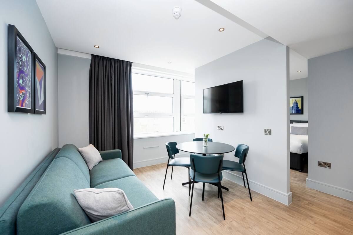 Un salon moderne avec un canapé de couleur turquoise, une table de salle à manger ronde entourée de chaises, un téléviseur à écran plat fixé au mur et une grande fenêtre agrémentée de rideaux.