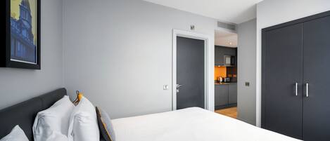 A modern hotel room with a large bed, a dark wardrobe, and a kitchen area visible through an open door.
