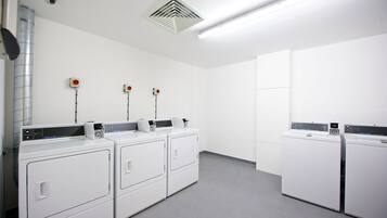 Un lavadero con cuatro lavadoras y secadoras de carga frontal blancas, un sistema de ventilación y tres detectores de humo en la pared.