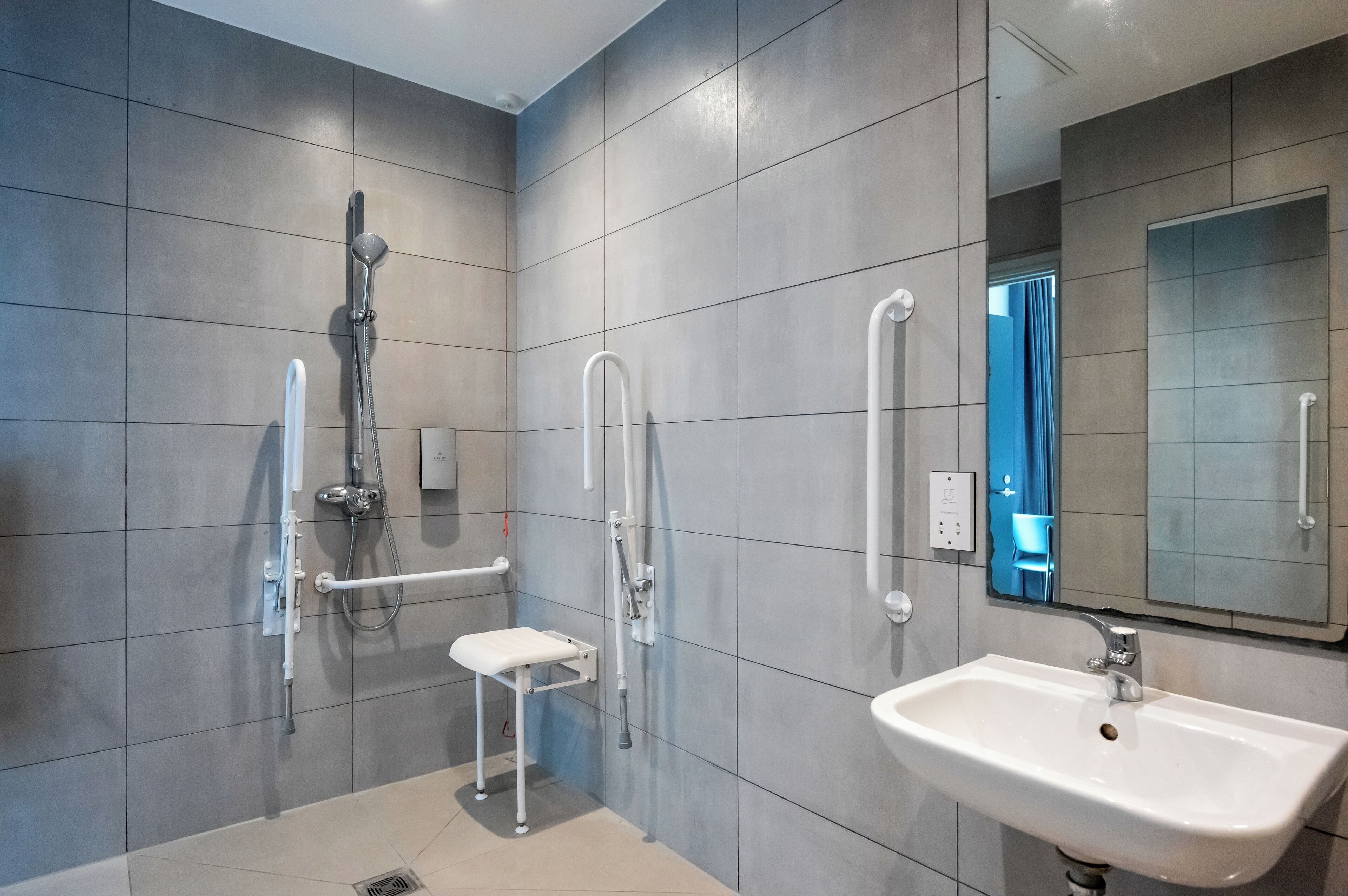 Un bagno moderno con piastrelle grigie, un lavandino bianco, un soffione doccia a mano e un piccolo sgabello bianco.