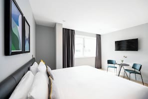 Quarto de hotel moderno com uma cama grande, uma TV presa à parede e um espaço de estar com duas cadeiras e uma mesinha.