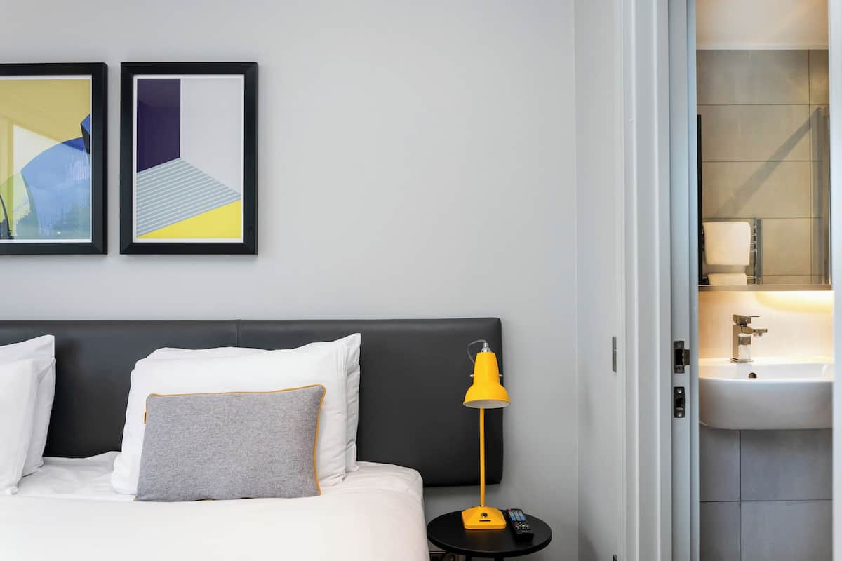 A hotel room with a bed, a nightstand with a lamp, a framed artwork, and a bathroom with a sink.