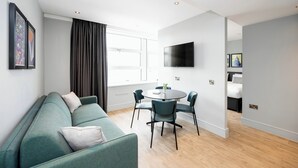 Een moderne woonkamer met een groene bank, een ronde eettafel met stoelen, een flatscreen televisie aan de muur en een groot raam met gordijnen.