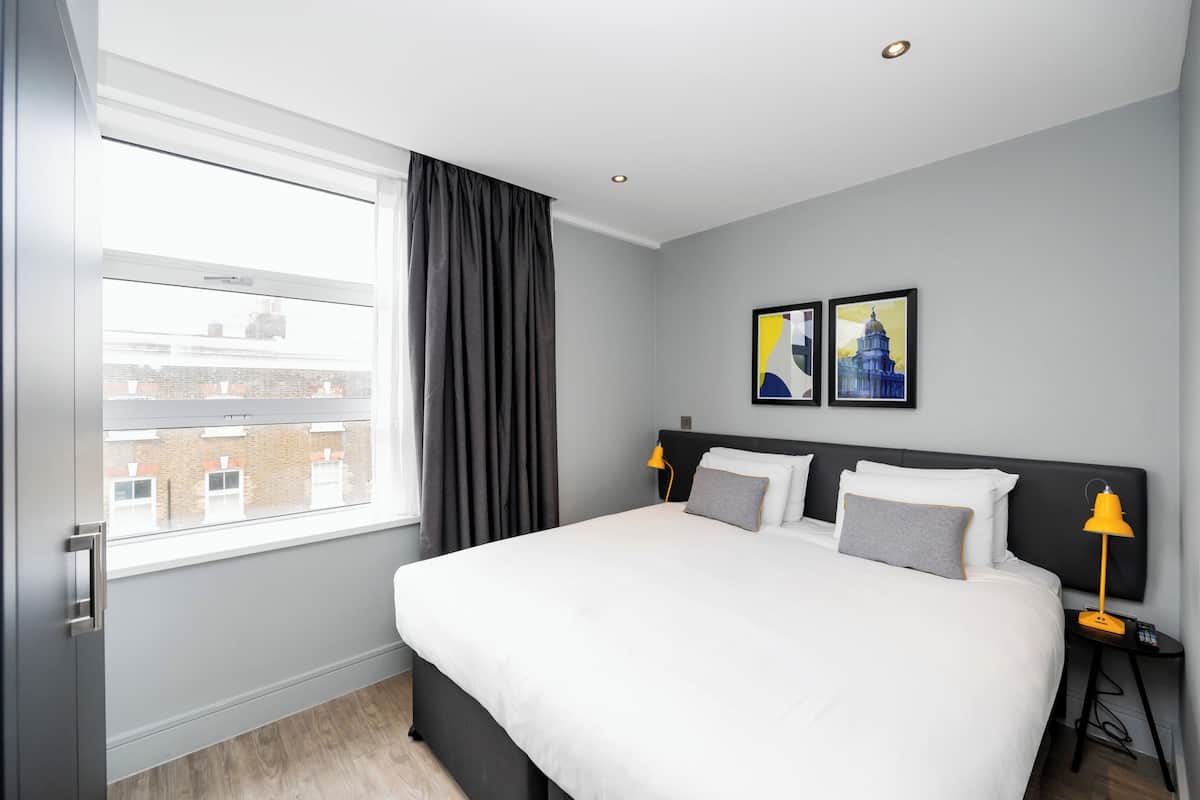 A modern hotel room with a large bed, a window with curtains, a nightstand with a lamp, and two framed pictures on the wall.