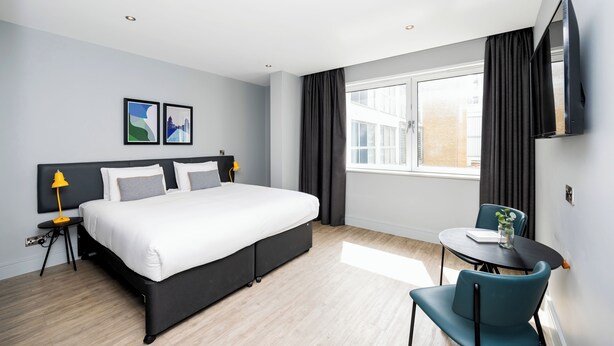 Een moderne hotelkamer met een groot bed, een klein tafeltje met stoelen, een aan de muur bevestigde televisie en een raam met gordijnen.