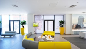 Een moderne lobby met geel en grijs zitmeubilair, grote ramen en een digitaal scherm.