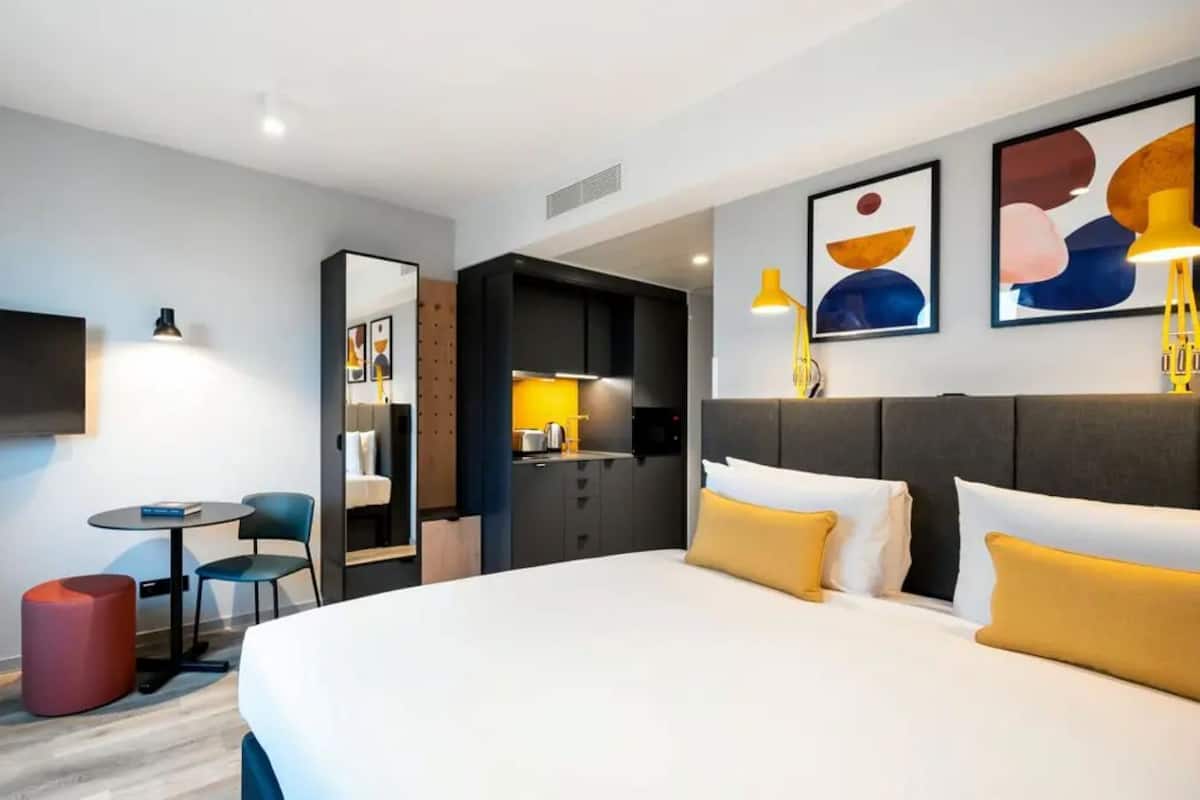 A hotel room with a bed, a desk, a chair, a small table, and a TV.