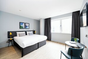 Ein modernes Hotelzimmer mit einem großen Bett, einem kleinen runden Tisch mit einem Stuhl, einem an der Wand befestigten Fernseher und einem Fenster mit Vorhängen.