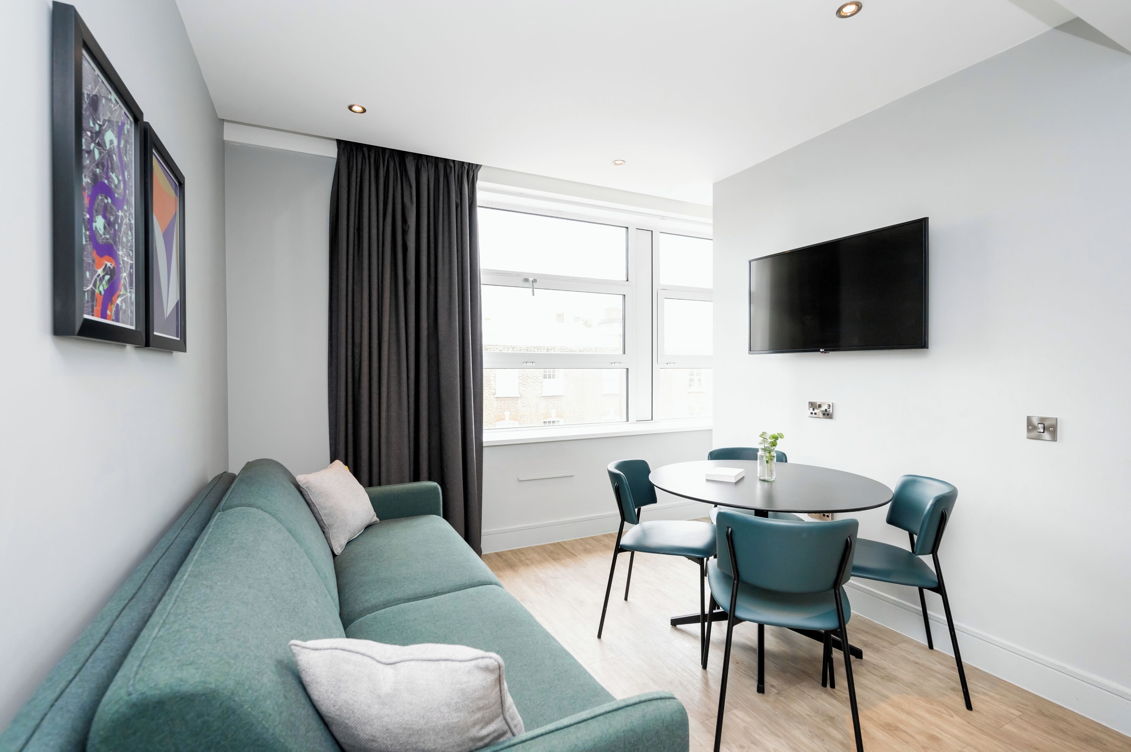Una sala de estar moderna con un sofá verde, una mesa de comedor redonda con sillas, un televisor de pantalla plana montado en la pared y ventanas grandes con cortinas.