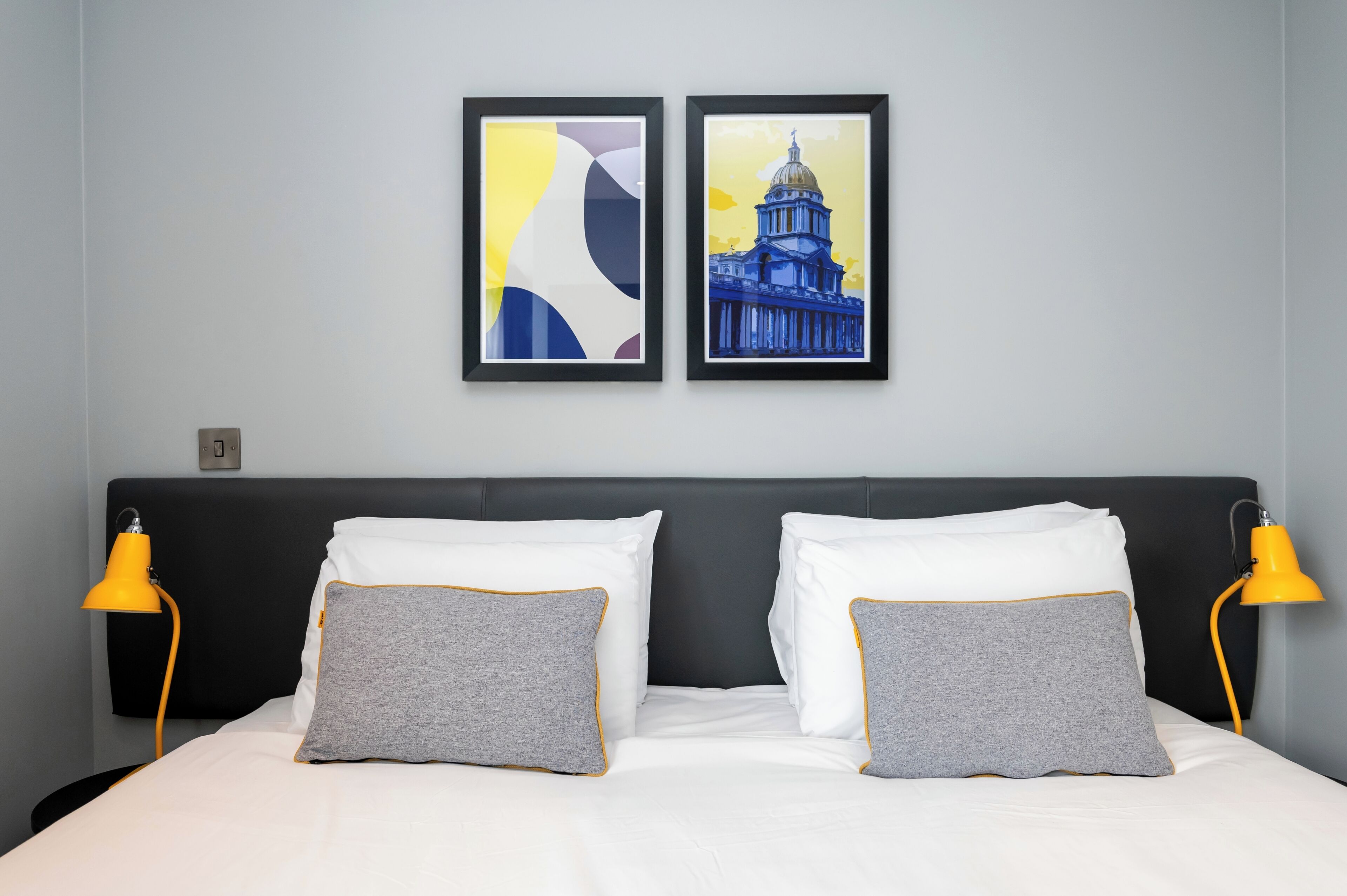 A hotel room with a bed, two pillows, two bedside lamps, and two framed pictures on the wall.