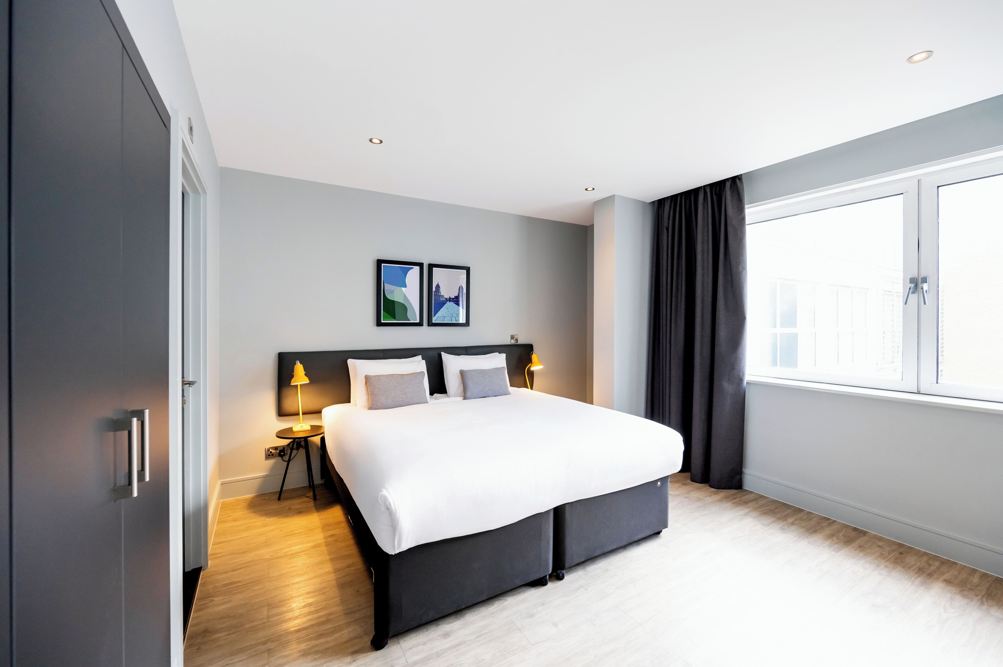 A modern hotel room with a large bed, two bedside tables with lamps, a window with curtains, and two framed pictures on the wall.