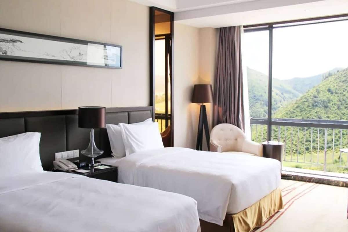 Une chambre d’hôtel avec deux lits, une table de chevet avec une lampe, une chaise et une grande fenêtre donnant sur un paysage montagneux.