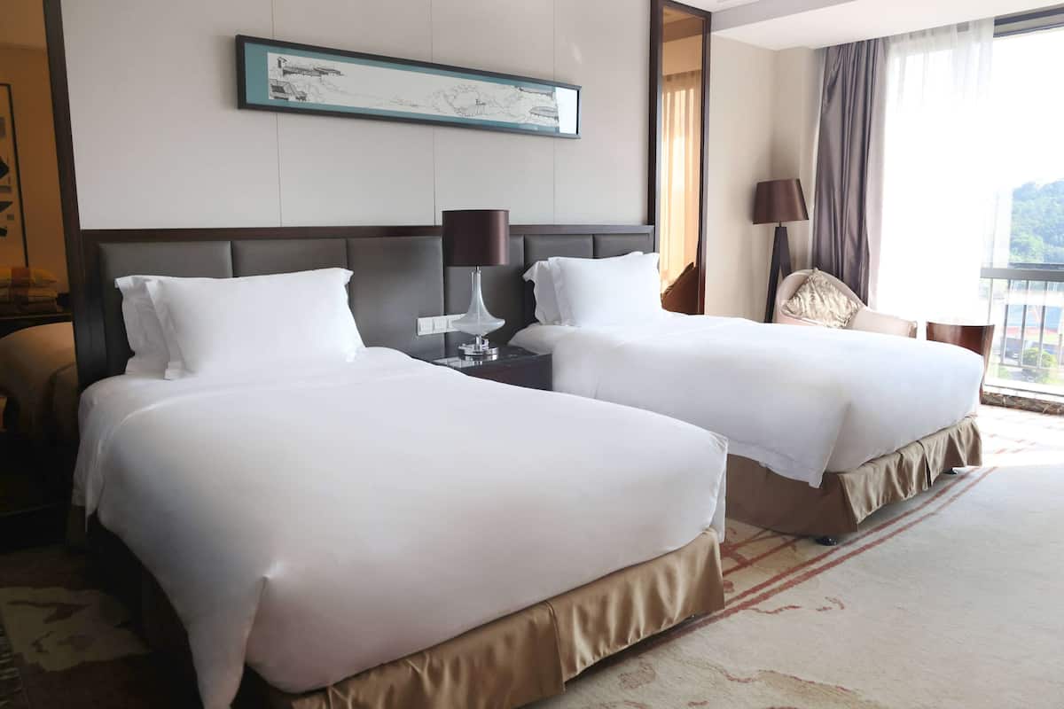 Une chambre d’hôtel avec deux grands lits, une table de chevet avec une lampe, une chaise et une grande fenêtre avec des rideaux.