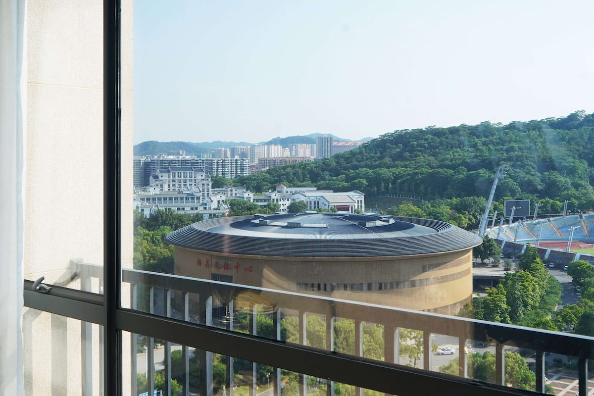 Une vue depuis une fenêtre donnant sur un paysage urbain, avec un bâtiment circulaire imposant et un stade bien visibles.