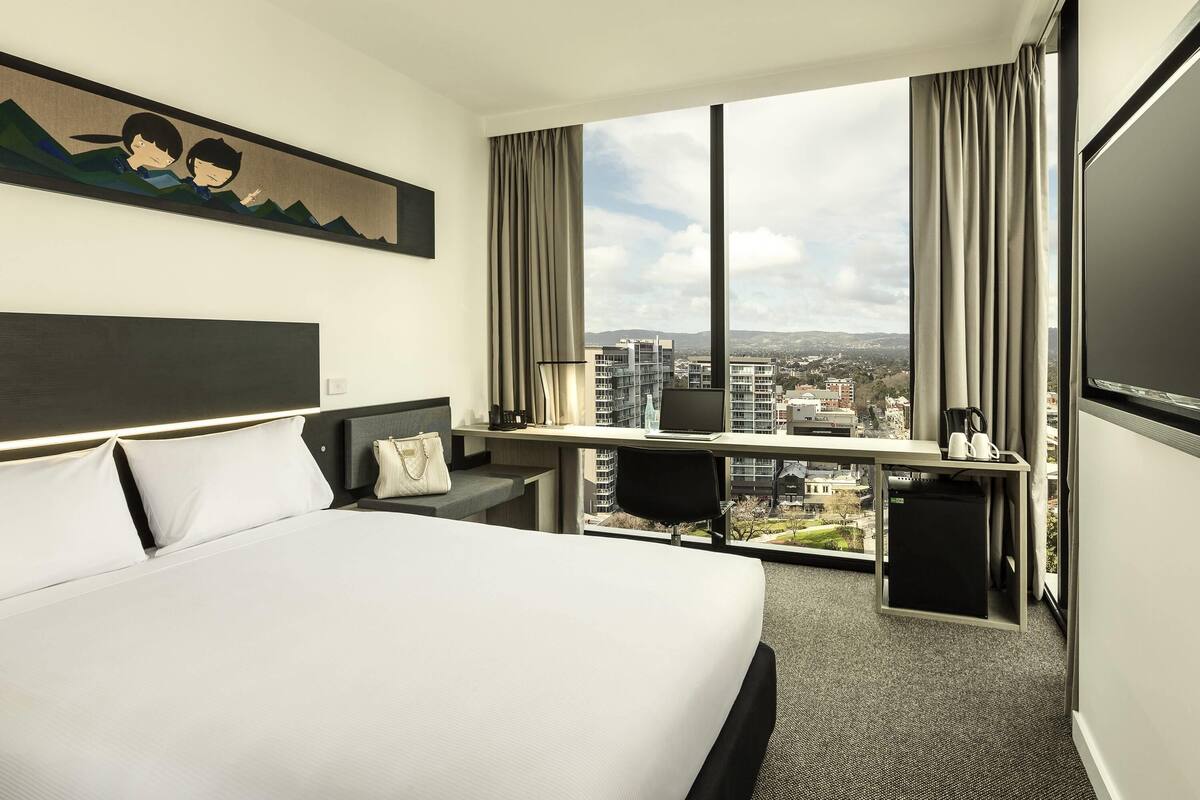 A hotel room with a bed, desk, and large window with a city view.