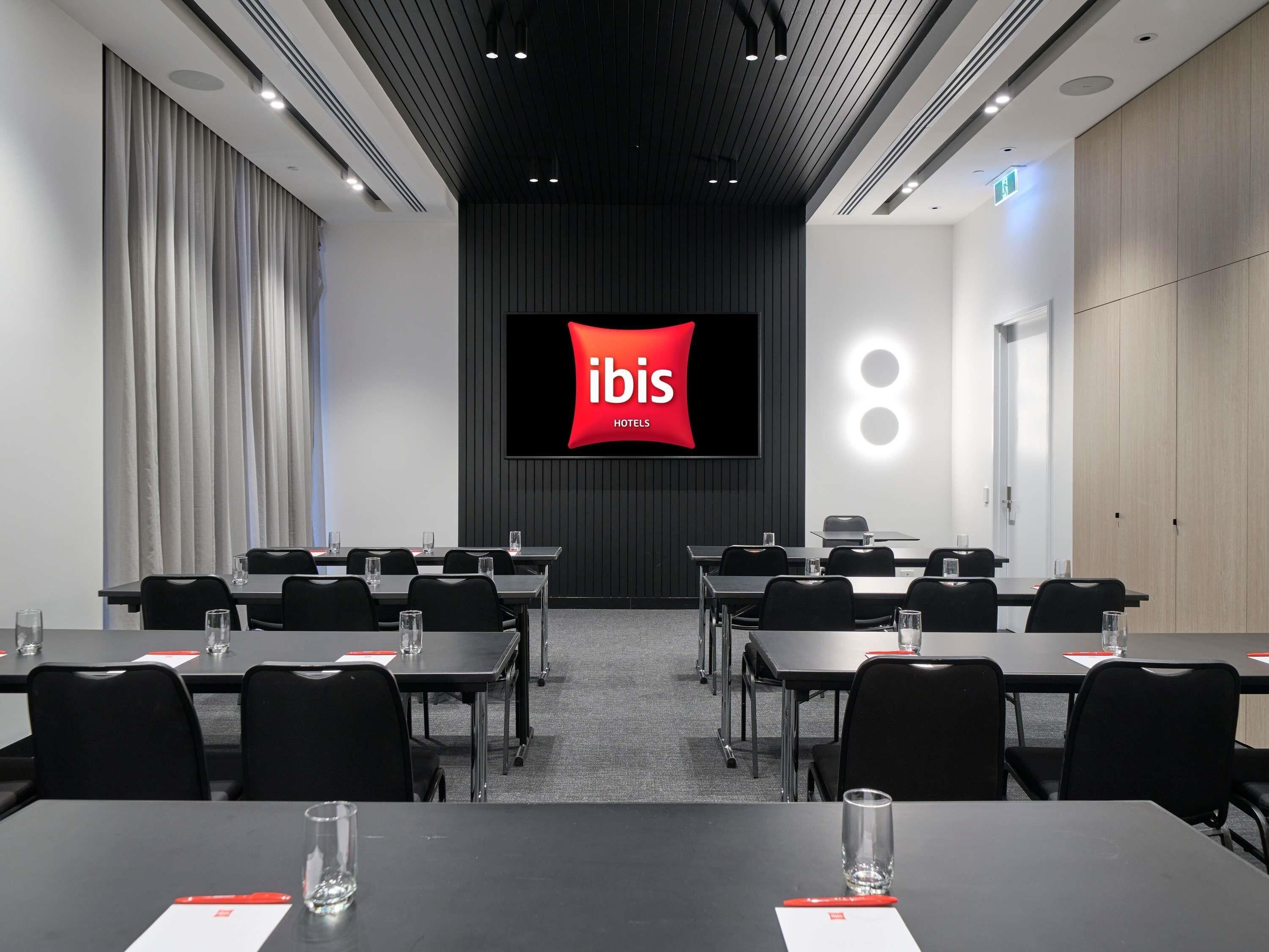 A conference room with a large screen displaying 'ibis HOTELS' logo, multiple tables with chairs, and a modern ceiling design.