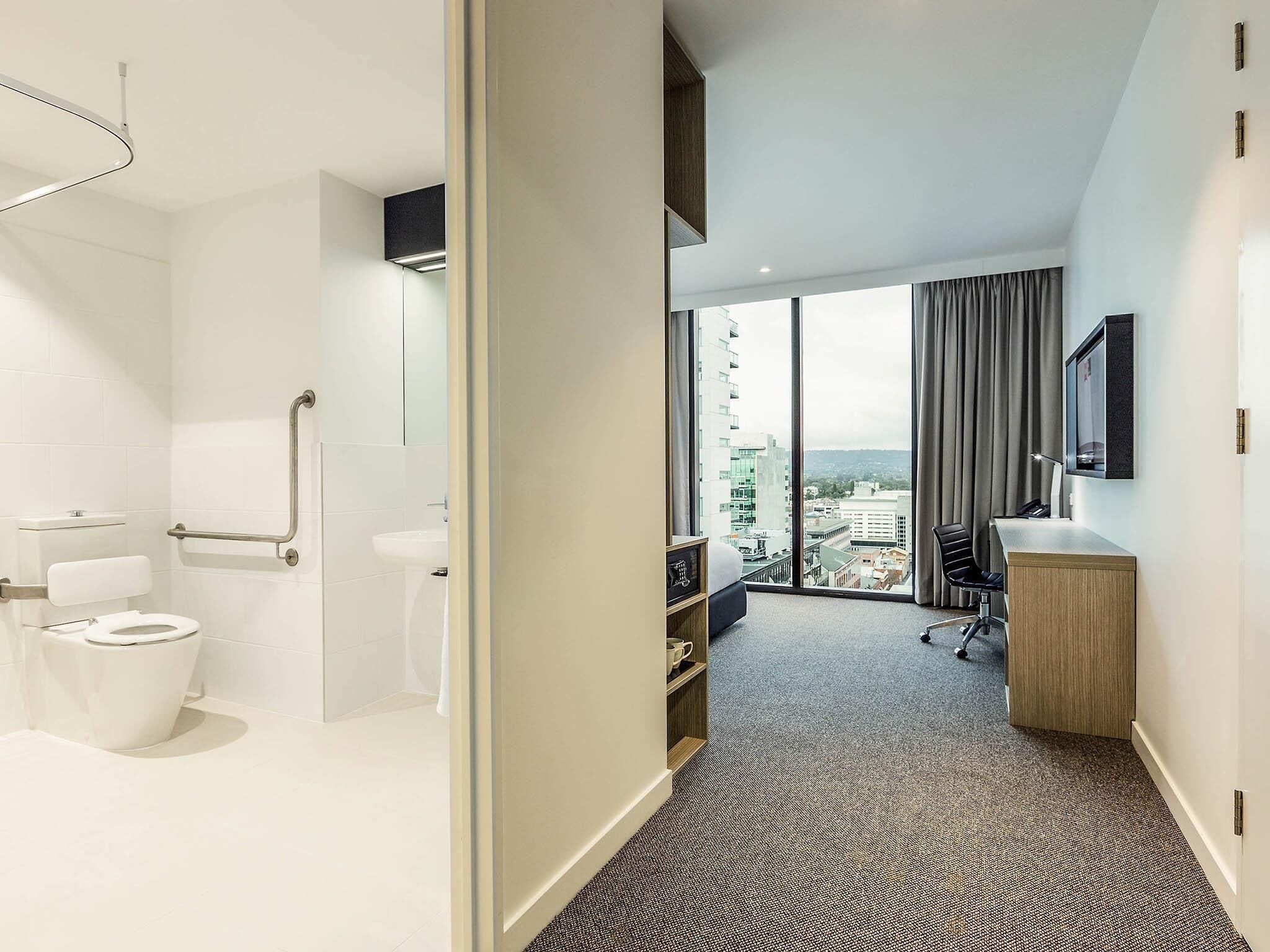 A modern hotel room with a bathroom, a large window with a city view, a desk, and a chair.