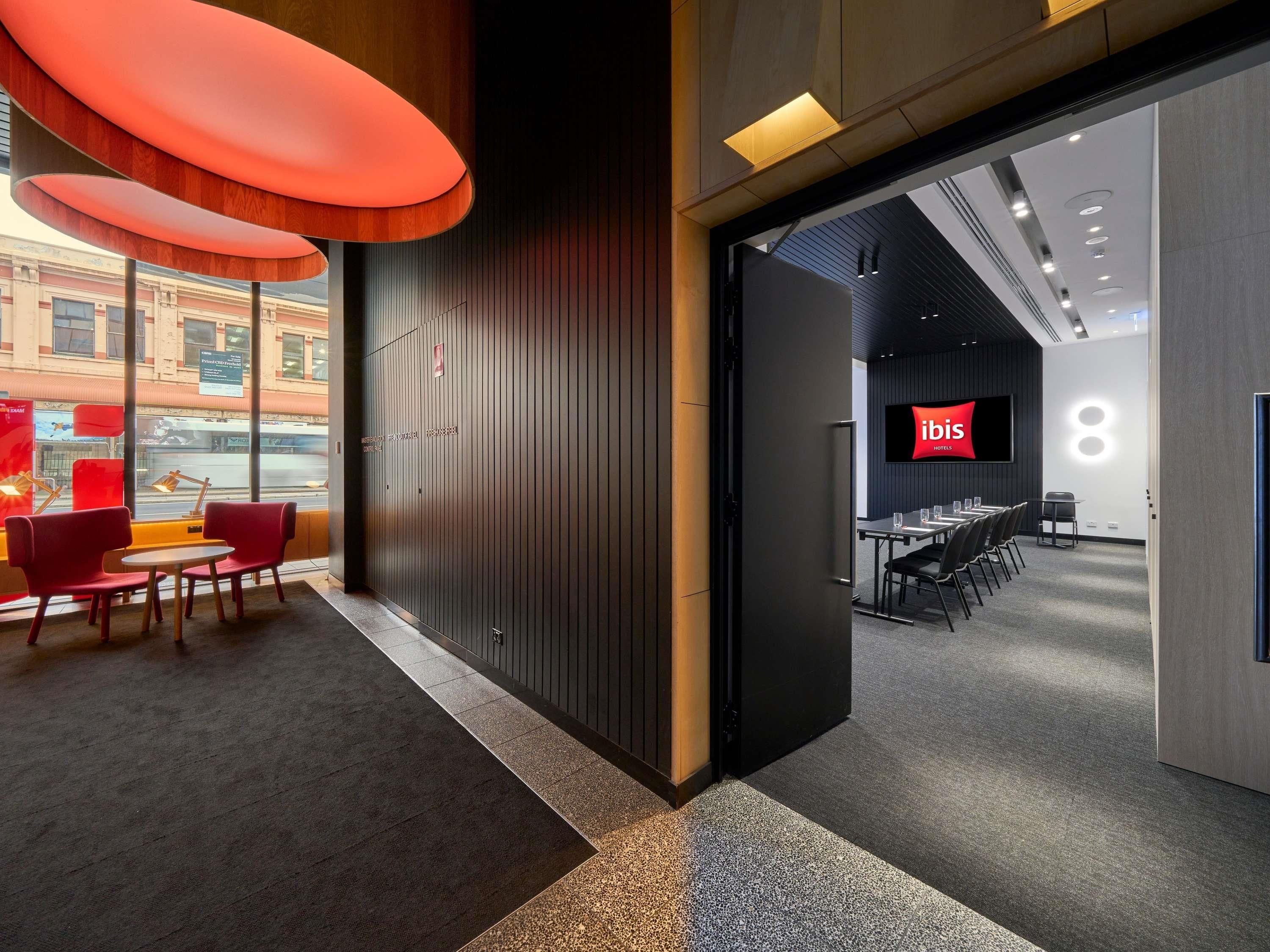 Un hall de hotel con diseño moderno, que incluye una sala de conferencias con mesas y sillas, una pantalla grande que muestra el nombre del hotel y vistas a la calle.