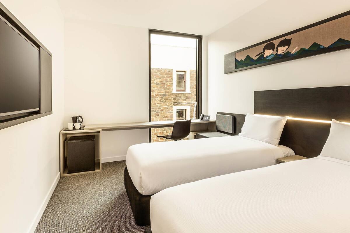 A hotel room with two beds, a TV, a desk with a chair, a small table with a coffee maker, and a window with a view of a stone building.