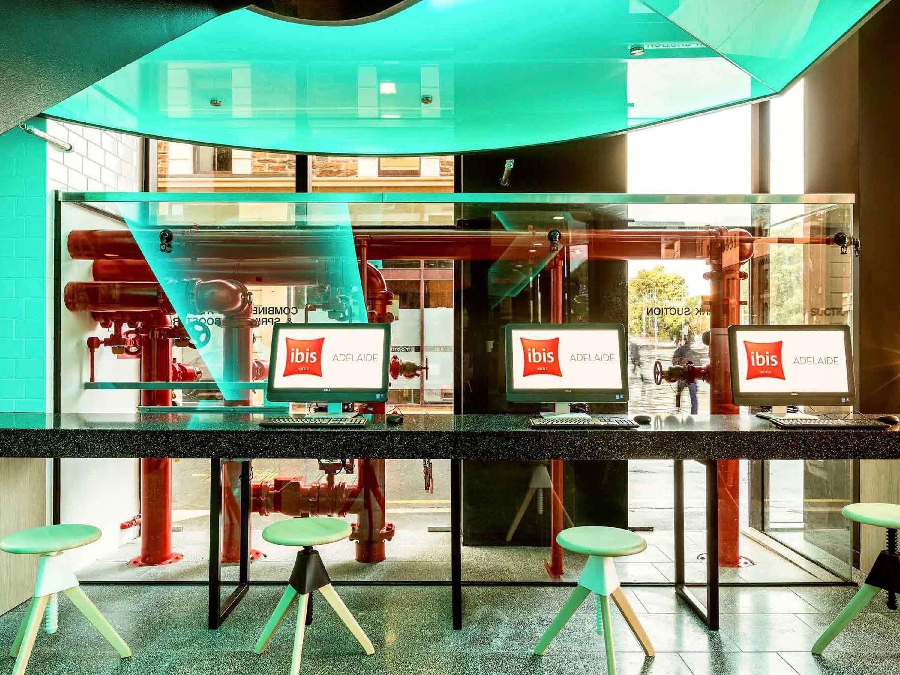 A modern hotel lobby with glass partitions, computers displaying the ibis logo, and a view of outdoor pipes.