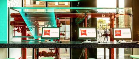 A modern hotel lobby with glass partitions, computers displaying the ibis logo, and a view of outdoor pipes.