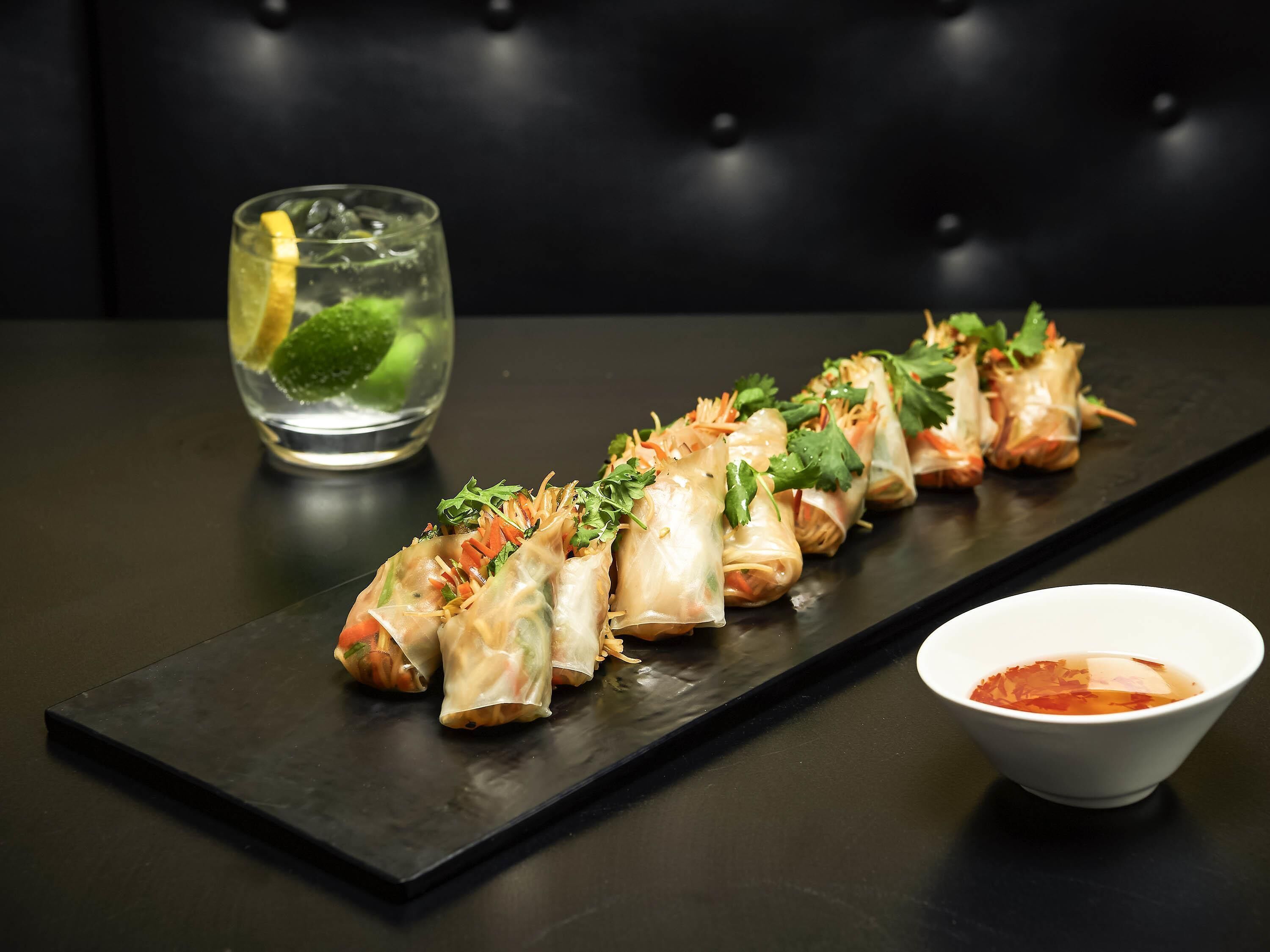 Una mesa con una fila de rollitos de primavera, un vaso de agua con una rodaja de lima y un tazón de salsa.