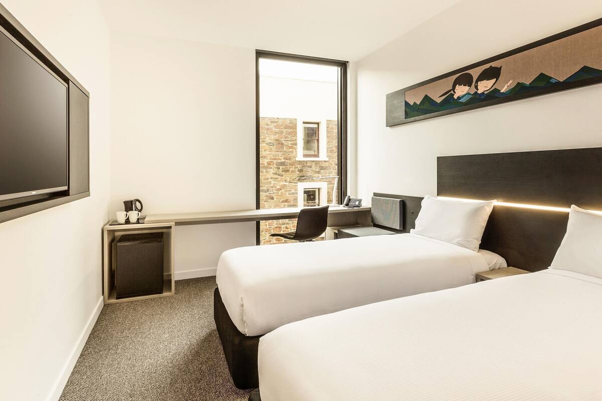 Habitación de hotel con dos camas, televisión, escritorio con silla, una mesita con cafetera y un cuadro en la pared.