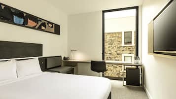 A hotel room with a bed, a desk with a lamp, a chair, a TV, and a stone wall visible through the window.