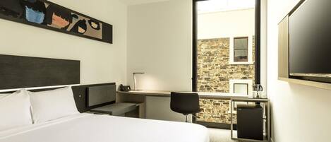 Habitación de hotel con cama, escritorio con lámpara, silla, televisor y pared de piedra visible a través de la ventana.