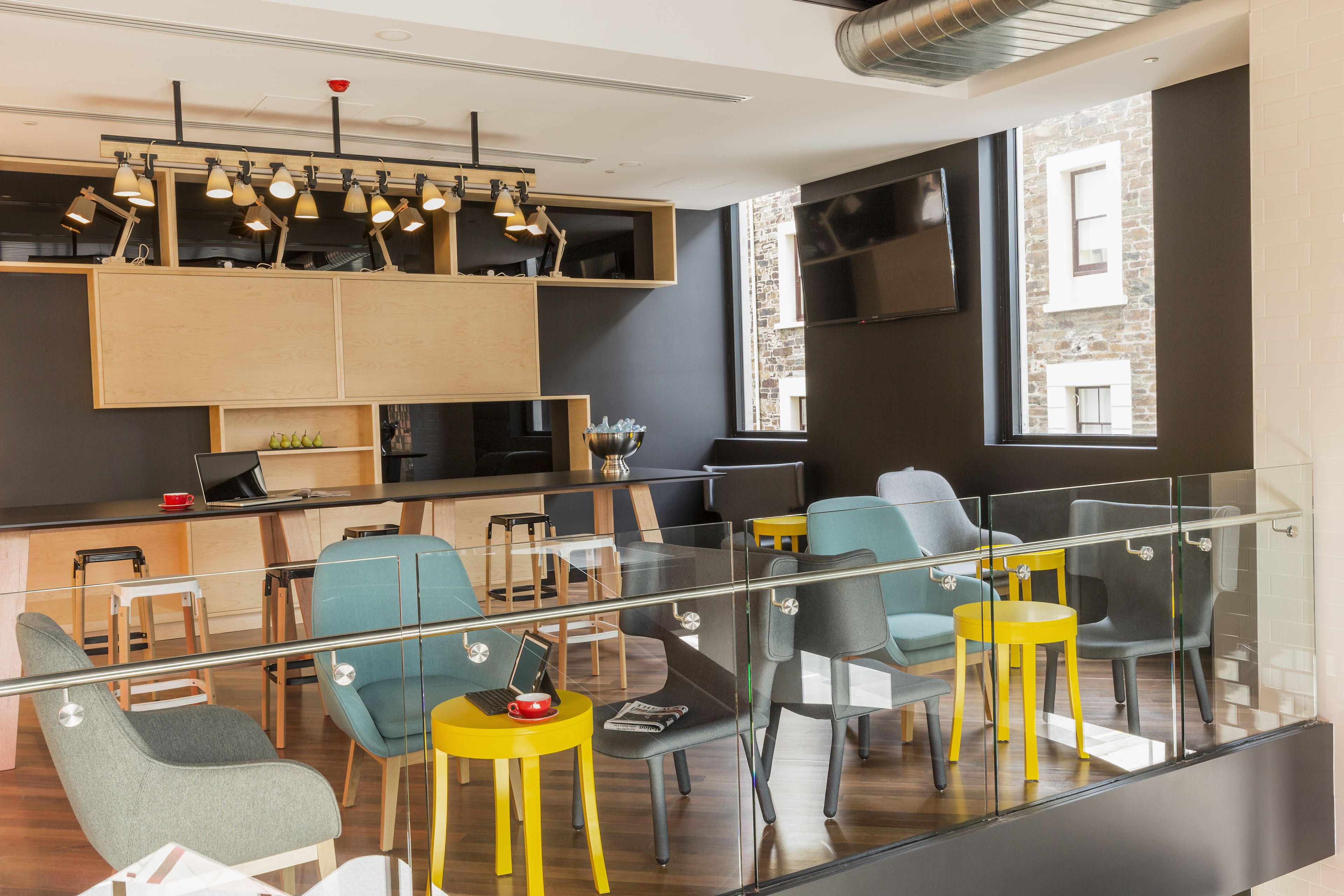 A modern lounge area with a glass railing, seating arrangements, and a kitchen area with a flat-screen TV.