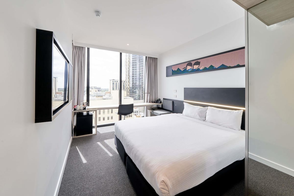 A hotel room with a large bed, a desk, a chair, a TV, and a city view.