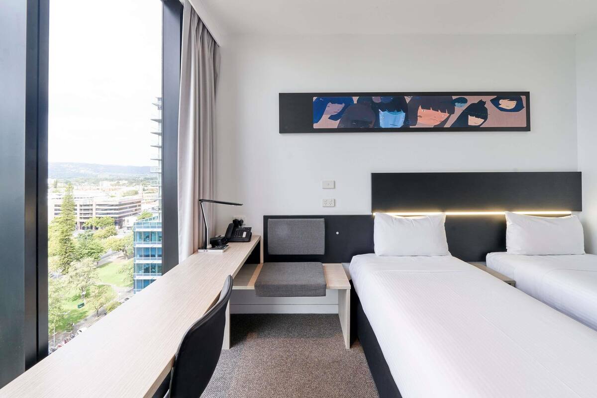 A hotel room with a desk, two beds, a view of the city, and a modern abstract painting.