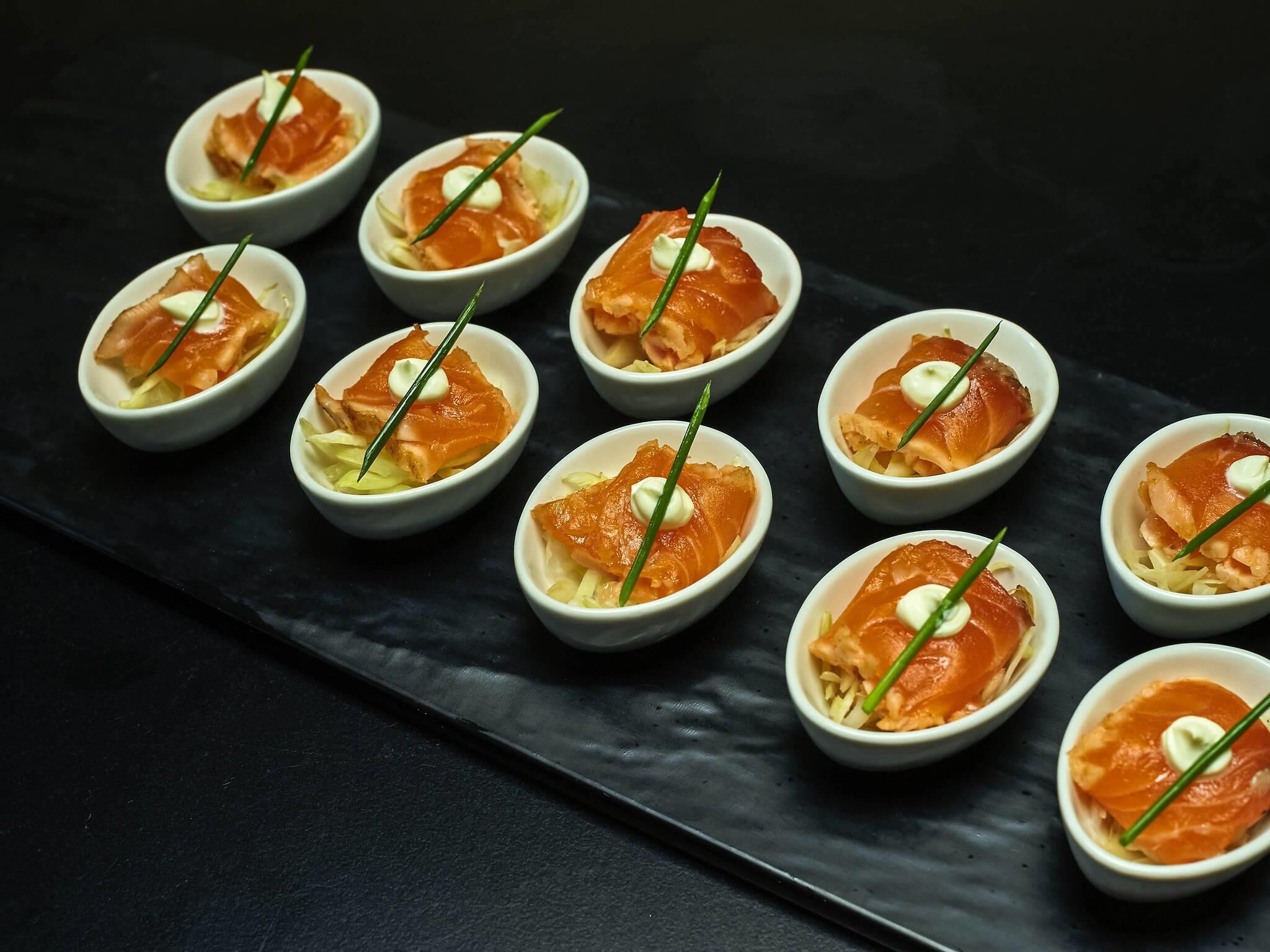 A series of small bowls containing a dish with smoked salmon and cream, garnished with chives, arranged on a dark slate serving board.
