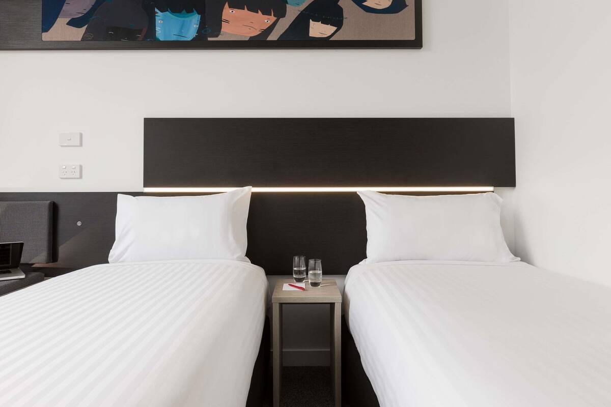 A hotel room with two beds, a nightstand with a glass of water, and a modern abstract painting above the bed.