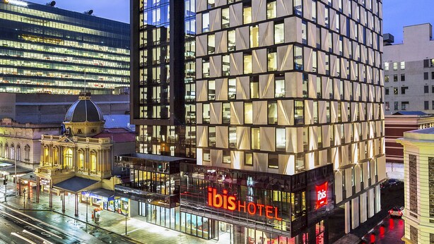 Un hôtel moderne doté d’une façade en verre, d’une enseigne « ibisHOTEL » bien visible et offrant une vue sur le paysage urbain environnant.