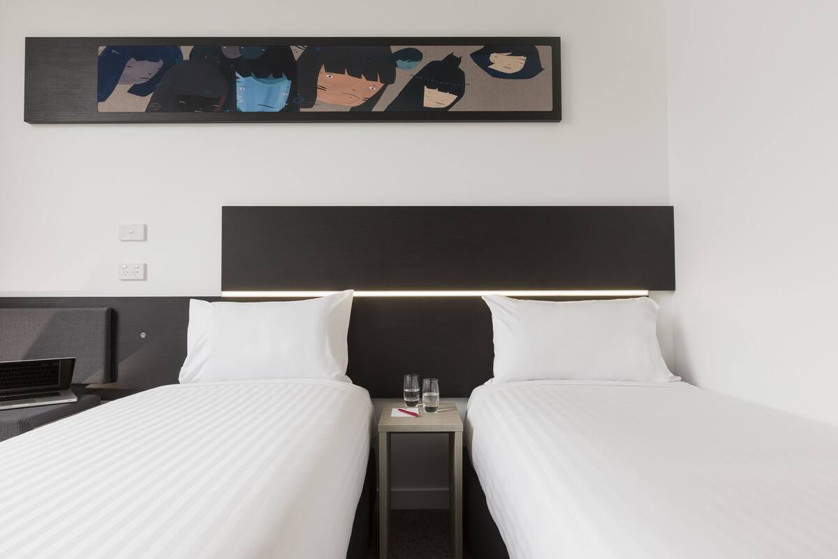 A hotel room with two beds, a bedside table with glasses and a book, and a wall-mounted artwork.