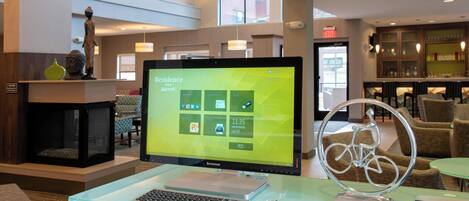 Eine Hotelhalle mit einer Rezeption, einem Computerbildschirm, der einen Desktop anzeigt, einem Glastisch mit einem Laptop, einer Fahrraddekoration und Sitzbereichen.